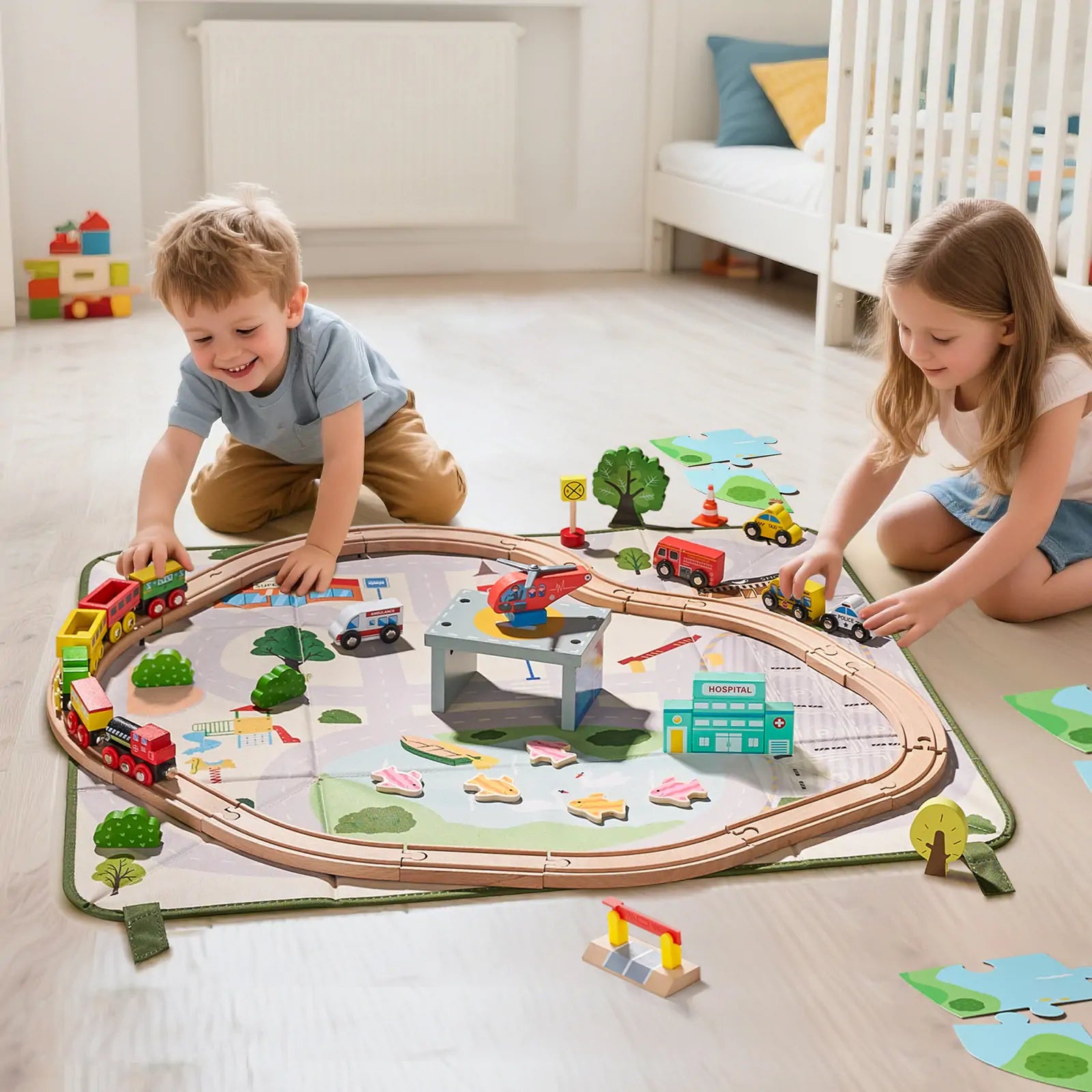 Two children play on the floor with the Tiny Land® 110-Piece Wooden Train Set, featuring tracks, vehicles, buildings, and signs atop a city play mat. The bright bedroom includes a crib and toys, and the set comes with a folding storage box.