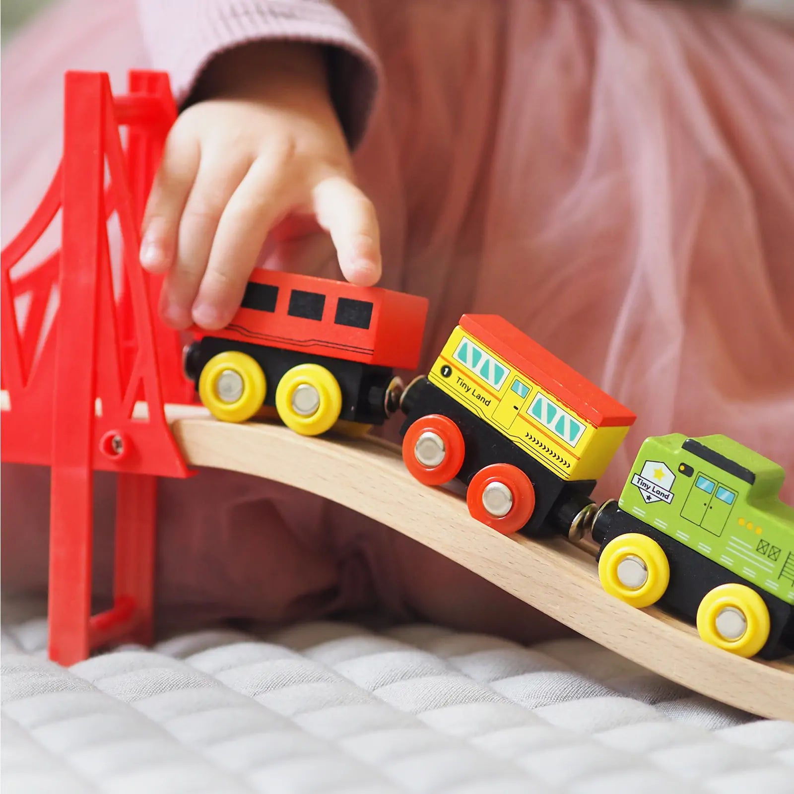 A child in pink pushes the Tiny Land® 110-Piece Wooden Train Set with Folding Storage Box over a red bridge on its track, with a city play mat in the background.