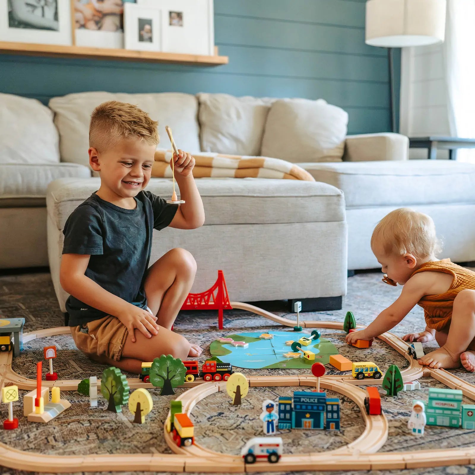 Two young children play with the Tiny Land® 110-Piece Wooden Train Set with Folding Storage Box. The older child smiles, holding a train piece, while the baby arranges tracks. In the background sits a beige sofa.