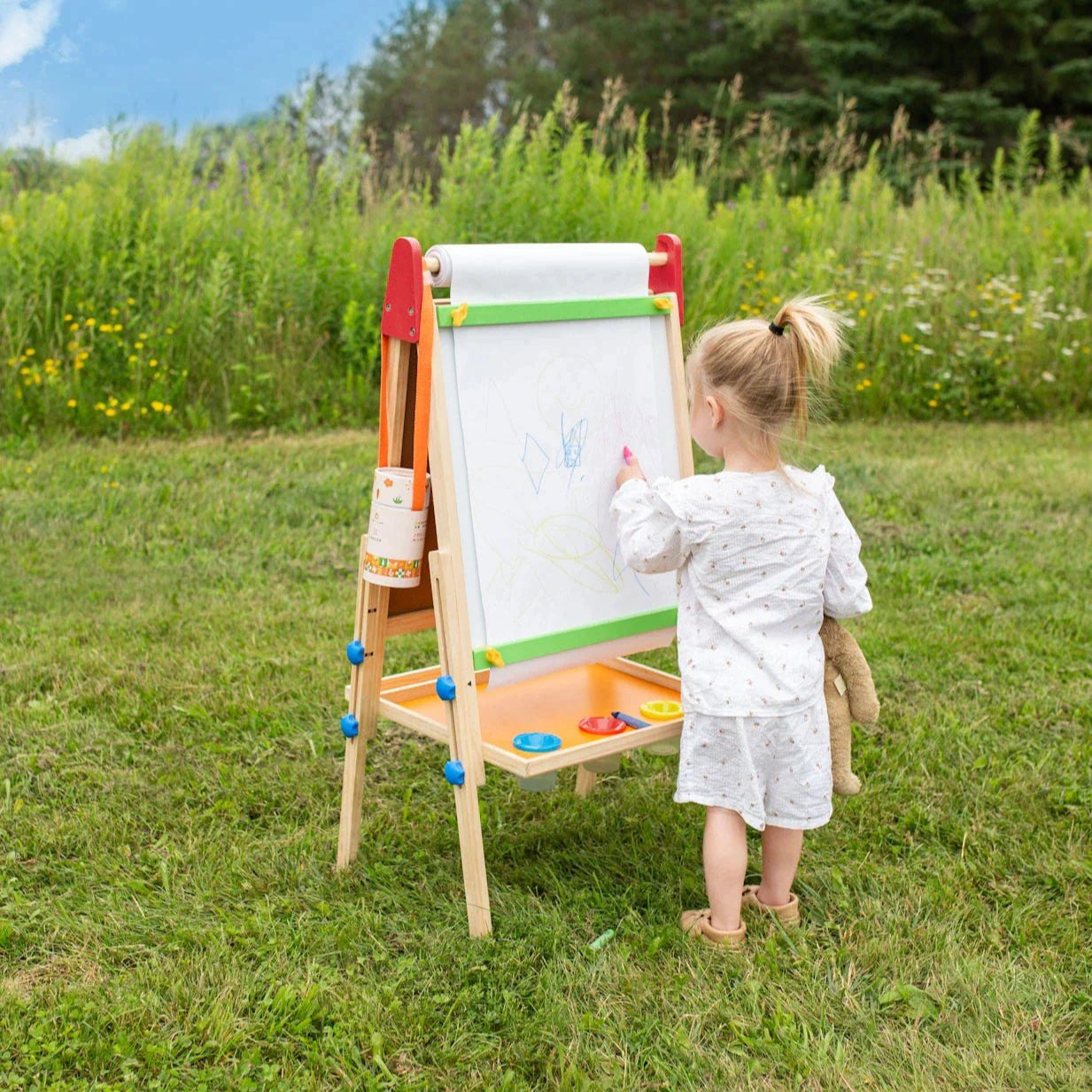 A young child in light pajamas holds a stuffed animal and draws on the Tiny Land® 3-in-1 Art Easel & Paper Roll Set in a grassy field, surrounded by wildflowers and trees, enjoying a peaceful moment of creativity.