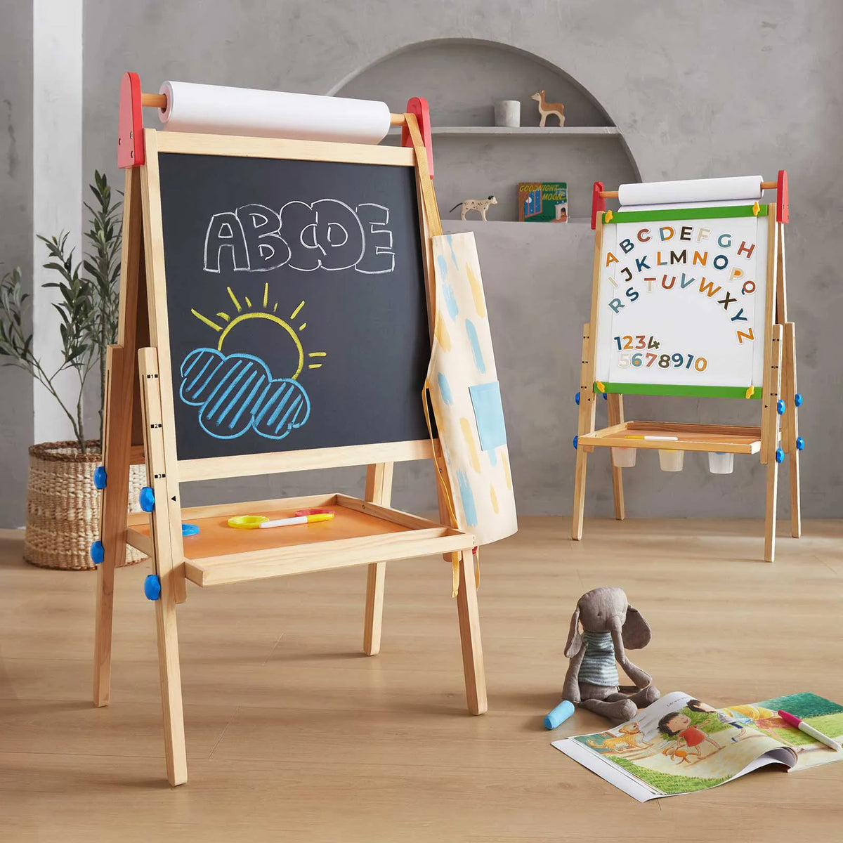 Two Tiny Land® 3-in-1 Art Easels for Kids stand in a playroom. The front, adjustable-height easel from Tiny Land features ABCDE and a sun drawn in chalk, while the back displays colorful magnetic letters on its whiteboard surface. A stuffed bunny and an open book rest on a rug in the foreground.
