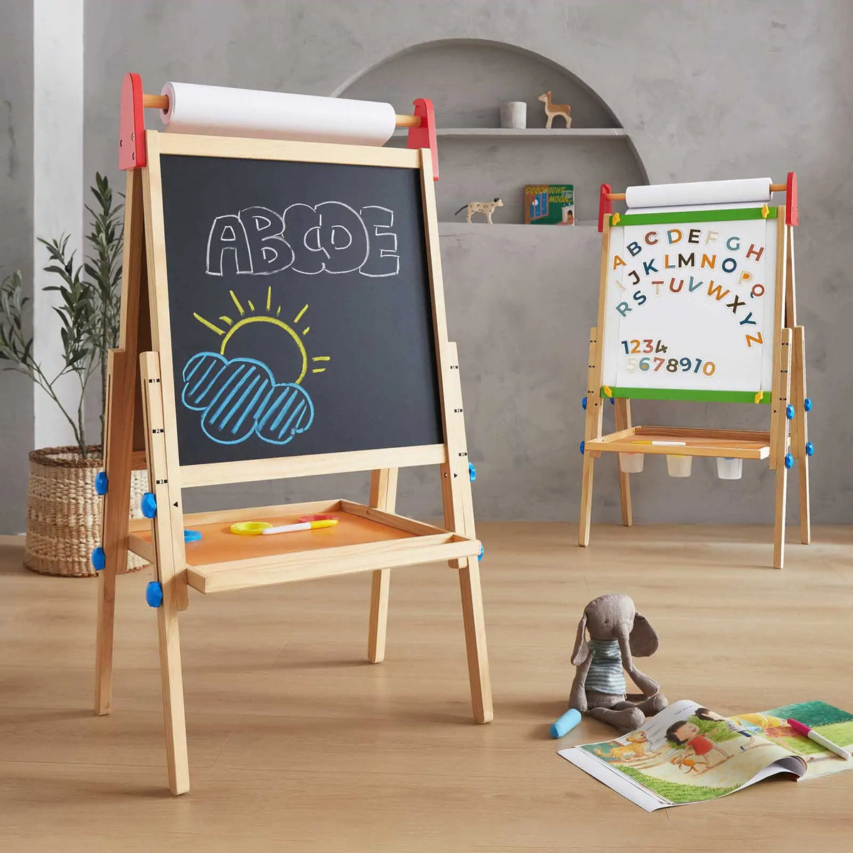 A children’s playroom features the Tiny Land® 3-in-1 Art Easel for Kids by Tiny Land; one side has a chalkboard with colorful drawings of ABCDE and a sun above clouds, while the other shows the alphabet. A plush bunny and drawing supplies sit on a nearby mat.