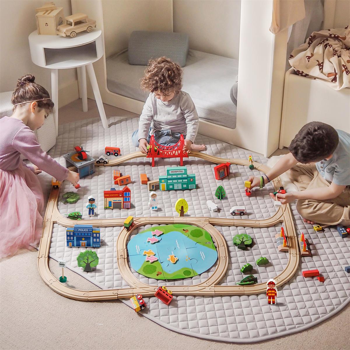 Three children play on a gray mat with the Tiny Land® Wooden Train Set 110 Pcs—compatible with all brands—surrounded by miniature buildings, vehicles, and trees in a cozy, softly lit room.|110 Pcs