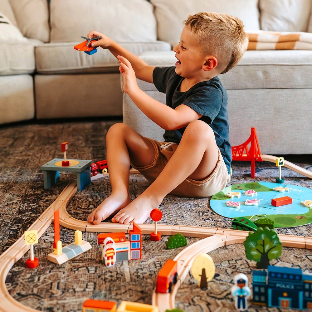 A young boy sits on the floor smiling and playing with the Tiny Land® Wooden Train Set 110 Pcs, compatible with all brands, along with a toy airplane, surrounded by colorful buildings and tracks in a cozy living room.