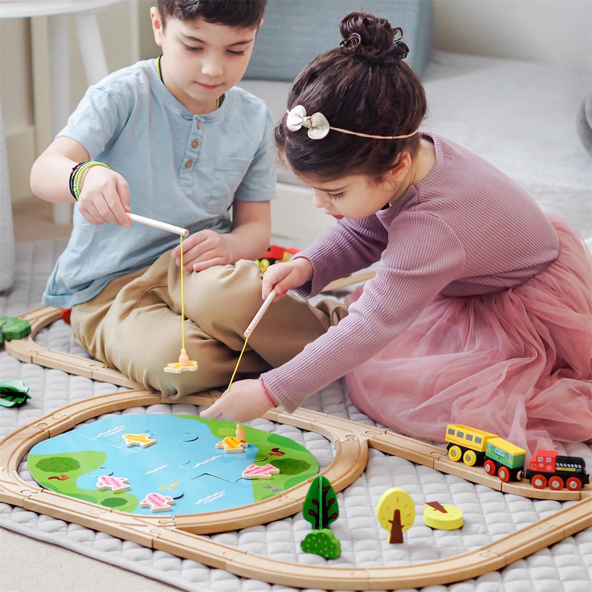 Two young children play on a soft mat with the Tiny Land® Wooden Train Set 110 Pcs. The girl in a pink dress uses a fishing rod, while the boy in blue helps by their Tiny Land train set, which is compatible with all major brands.