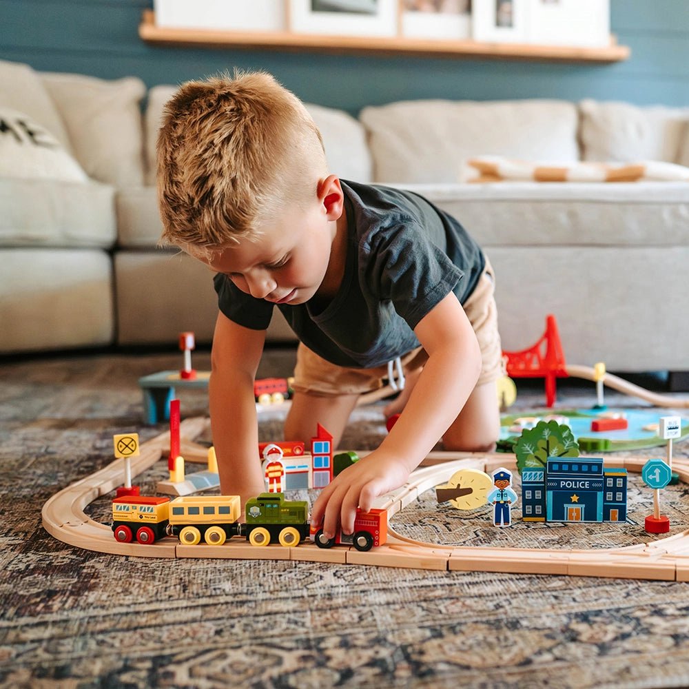A young boy plays on the floor with the Tiny Land® Wooden Train Set 110 Pcs, compatible with all brands, surrounded by miniature buildings, signs, and tracks in a cozy living room with a sofa in the background.