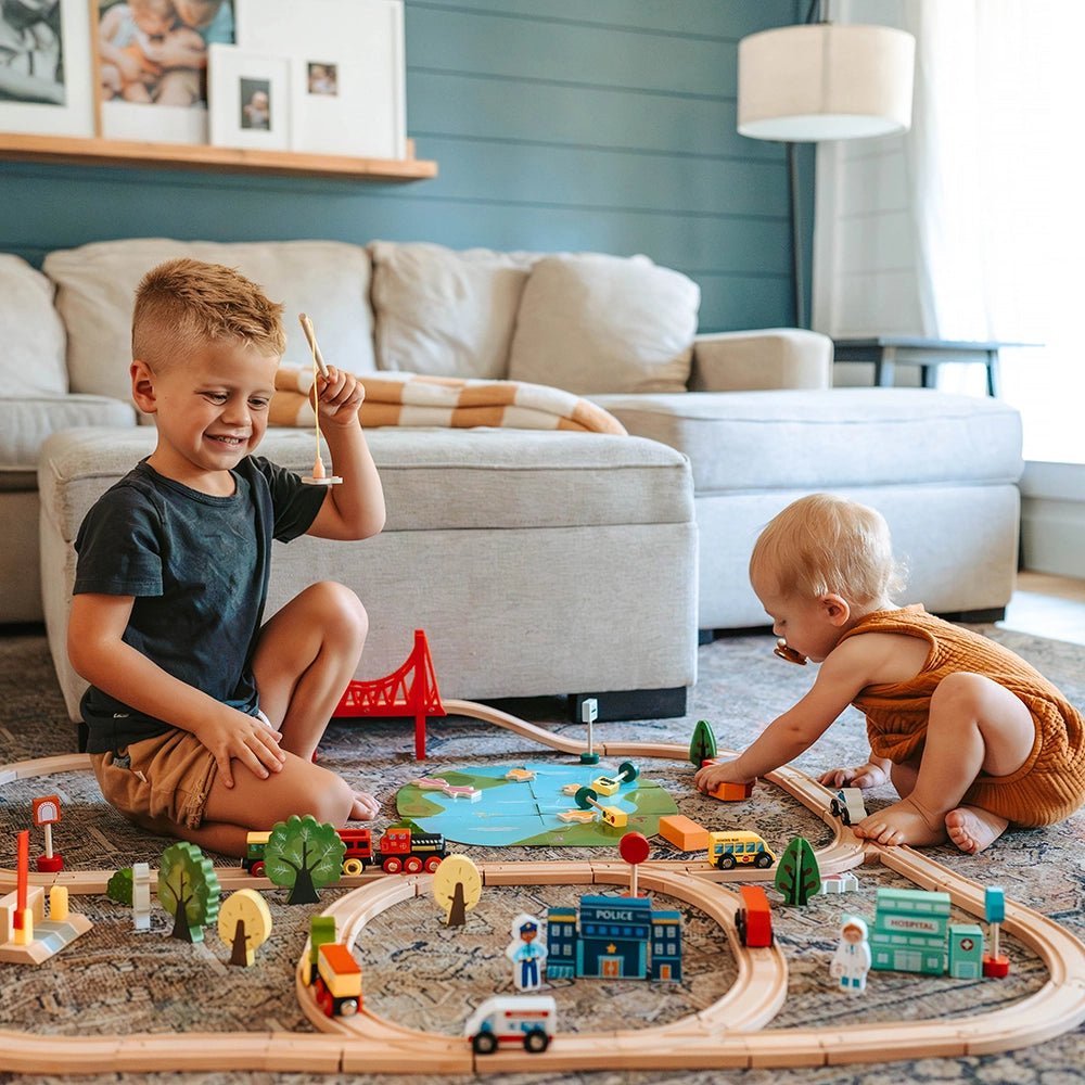 Two young children play on a light-colored rug with the Tiny Land® Wooden Train Set 110 Pcs. The older child smiles while holding a train, as the younger explores tracks and toys compatible with all brands from Tiny Land.