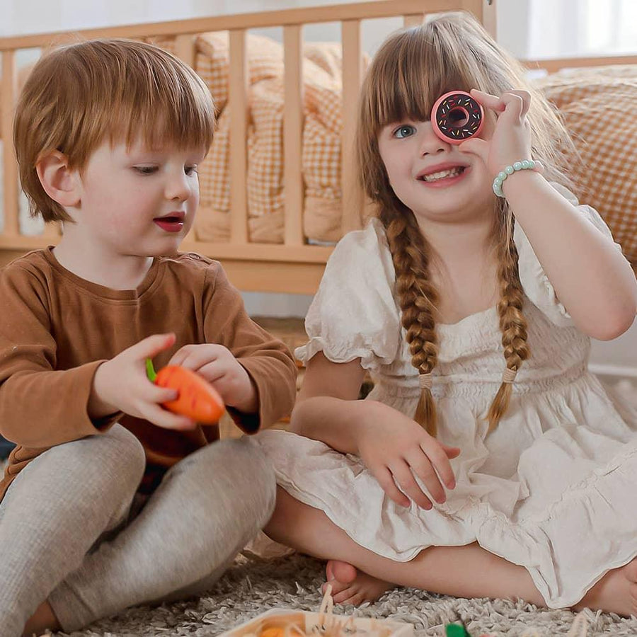 Two young children sit on a carpet with a crib behind them. The girl in white holds a Tiny Land® Wooden Food for Play Kitchen piece to her eye, while the boy in brown holds a toy carrot from the Tiny Land Play Food set, both having fun together.