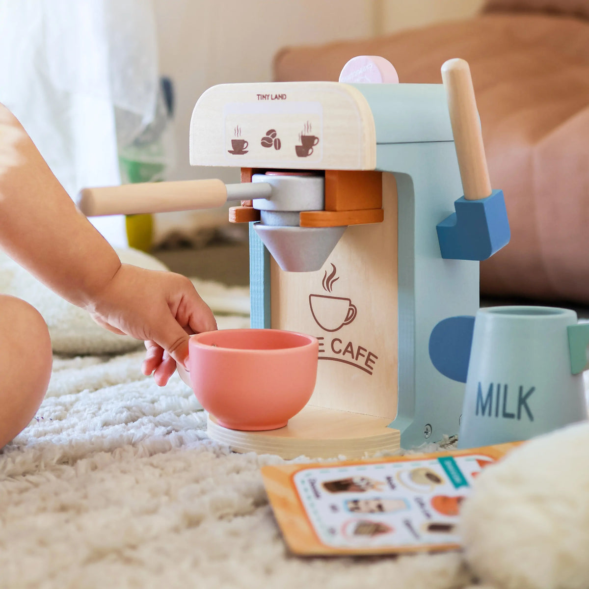 A babys hand reaches for a pink cup next to the Tiny Land® Play Kitchen Accessories wooden coffee maker, surrounded by kids’ kitchen toys and an illustrated menu on a soft carpeted floor.
