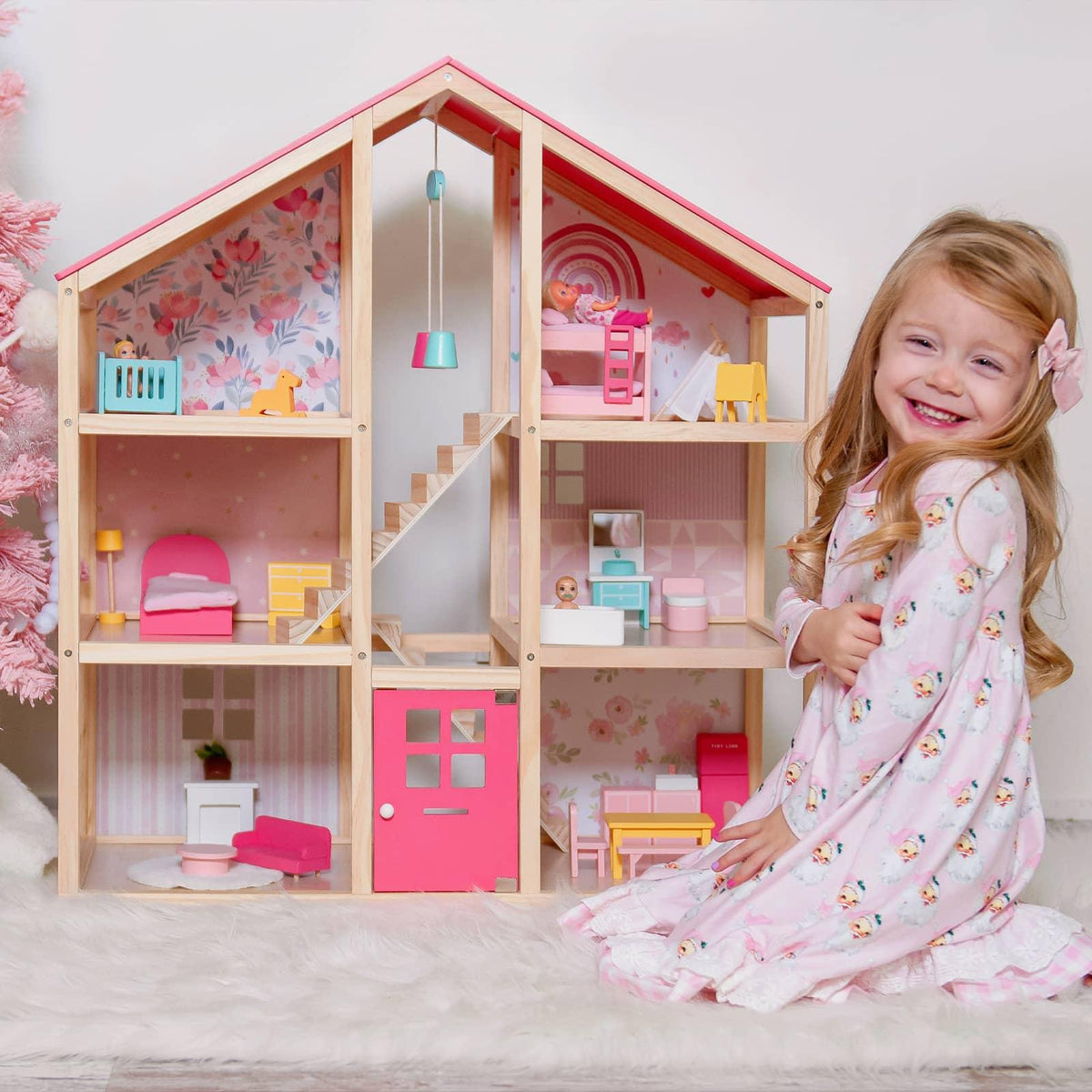A smiling girl in a pink floral nightgown sits by the Tiny Land Sweet Dreams Playset—Pink Dollhouse + Pink-White Playhouse, both decorated with colorful miniature furniture and wallpaper.|pink