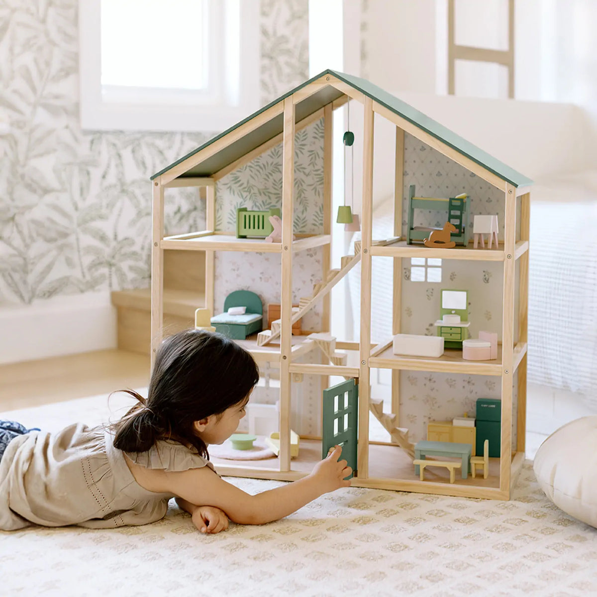 A young child plays on the floor, arranging miniature furniture in the Tiny Land® Sweetwood Evergreen Dollhouse without Dolls by Tiny Land. The room features light-colored walls and a patterned rug.