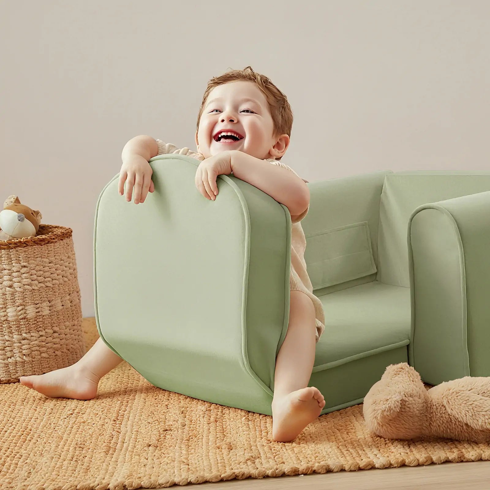 A smiling young child sits playfully on the Tiny Land® GentleSnug Kid Chair in Sage, surrounded by a woven basket, plush toy, and soft beige rug from Tiny Land in a cozy room.