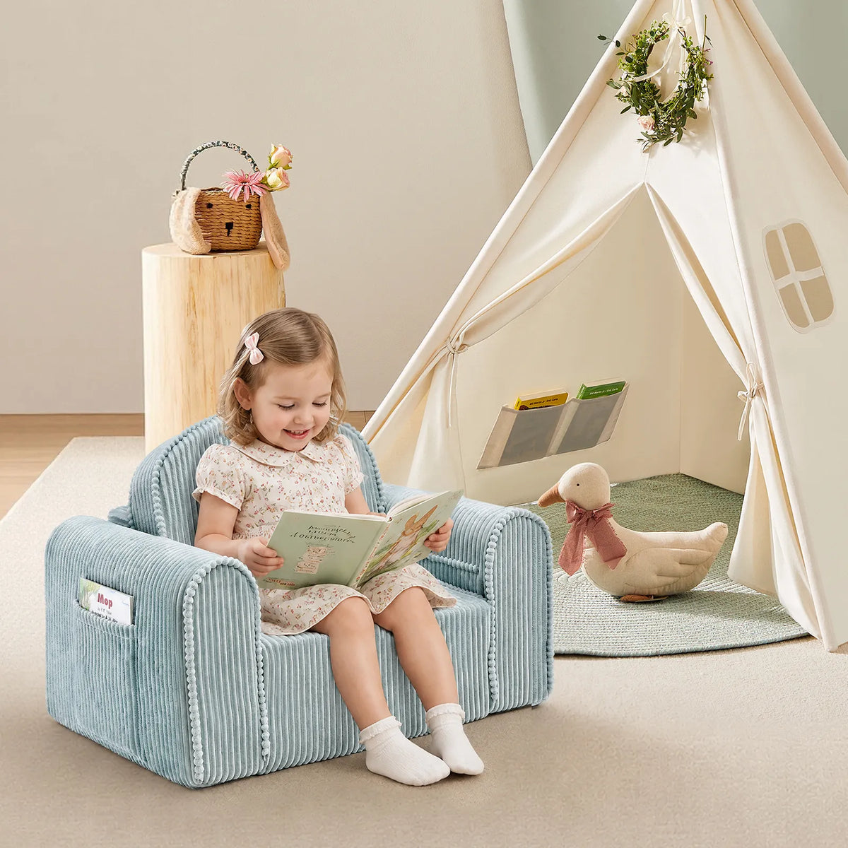 A young girl sits on the Tiny Land® Corduroy Lounger reading a book. Next to her is a basket on the table, a plush duck, and the Montessori-inspired Ultimate Reading Retreat Premium Teepee by Tiny Land INC, filled with books. The softly lit room feels cozy and inviting.|Soft Teal