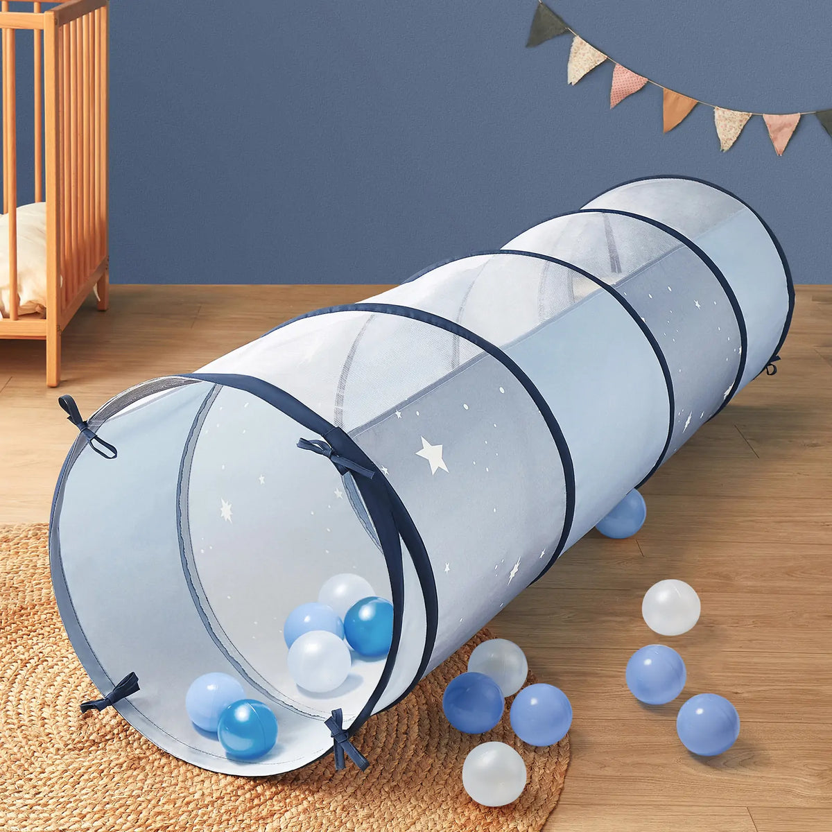 The Tiny Land® Adventure Play Tunnel - Blue by Tiny Land, featuring blue mesh and navy trim, rests on a wooden floor surrounded by blue and white balls in a playfully decorated kids' room with a crib, wall decor, and rug.