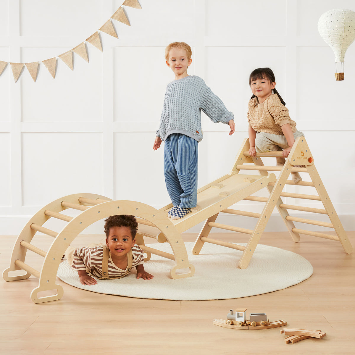 Three children enjoy the Tiny Land Safe Explorers Playset—one stands atop the natural wood climbing set, another climbs, and a third crawls beneath. The Urban Zoo Playmat and fun decor complete the bright, cozy playroom scene.