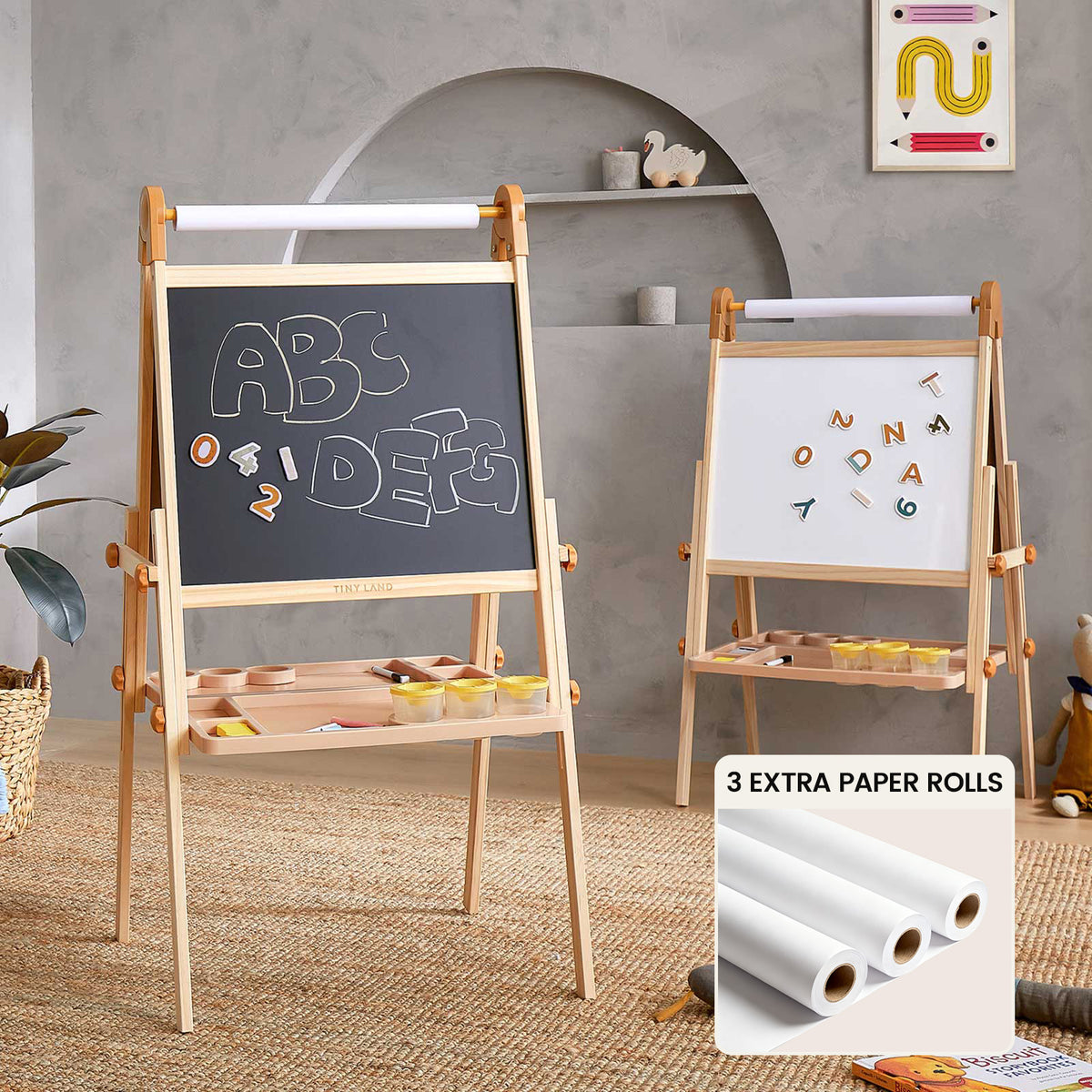 Two Tiny Land® Caramel Magnitales Easels are in a playroom—one with a magnetic blackboard and letters, the other a magnetic whiteboard. A Tiny Land kids art set with three extra paper rolls is shown in front.