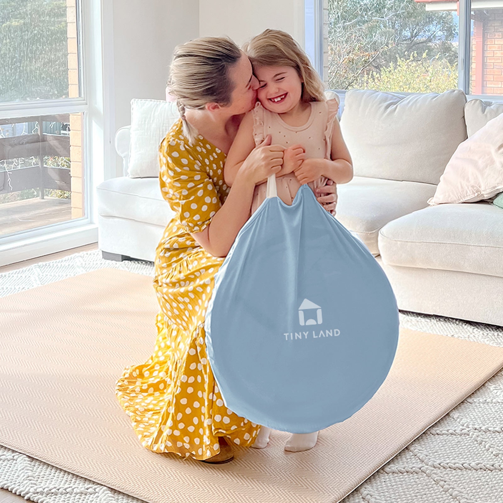 A smiling woman hugs a laughing girl holding the Tiny Land® Kids Tent – Starry Blue Indoor Tent for Toddlers beside a kids playhouse in a bright living room with a white sofa and large windows.