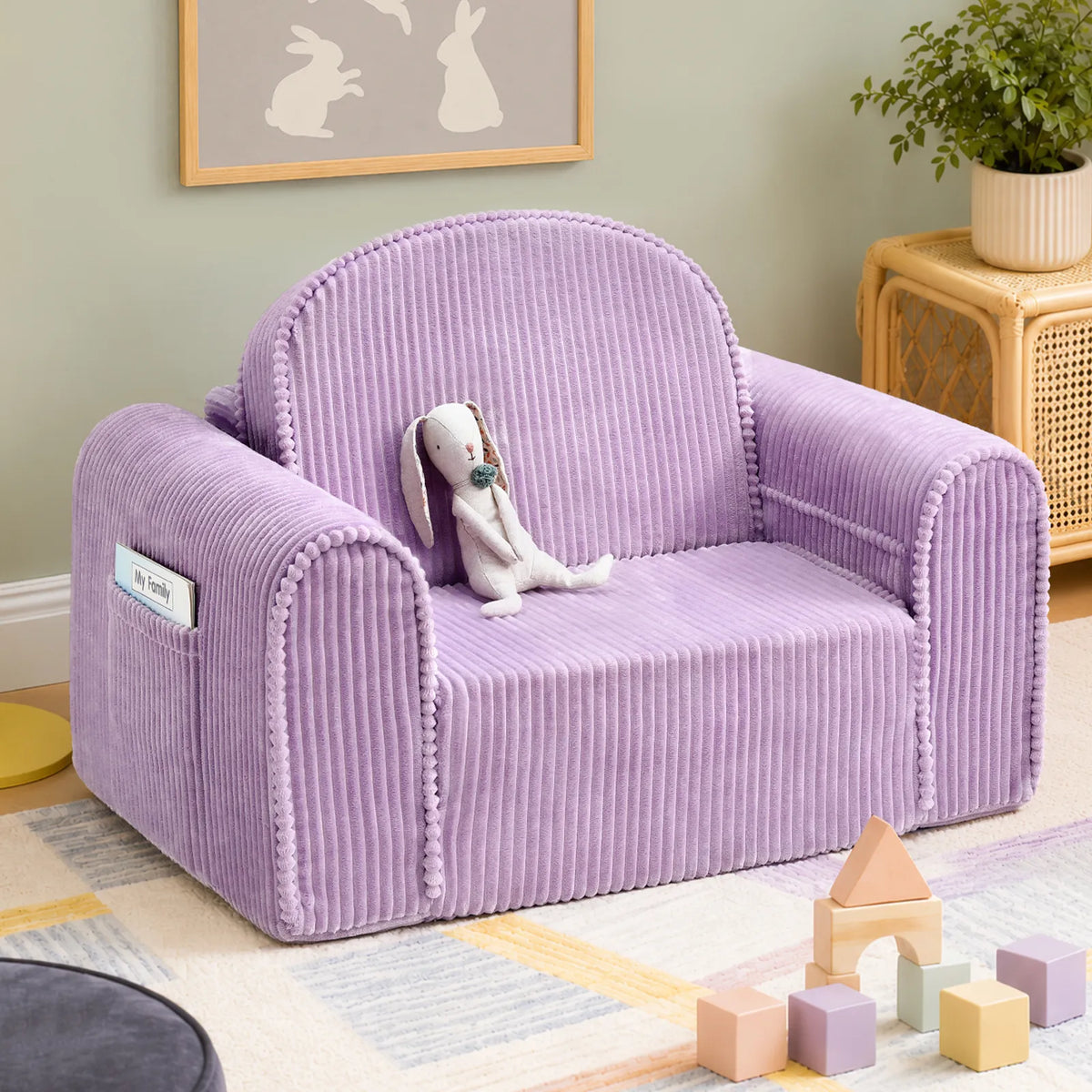 The Tiny Land® GentleSnug Corduroy Kid Chair by Tiny Land, in a soft lavender hue, holds a cuddly stuffed bunny in a cozy room. Wooden blocks and pastel toys are scattered on the floor, while a framed picture of rabbits hangs above, completing this charming corner of corduroy furniture.|Purple