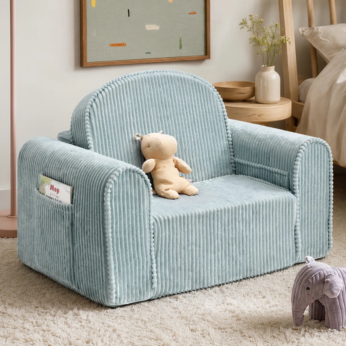 The Tiny Land® GentleSnug Corduroy Kid Chair in light blue serves as a charming kids lounger, adorned with a stuffed bear, and is set in a cozy room featuring a framed art piece, wooden furniture, and a plush elephant toy on a textured rug.