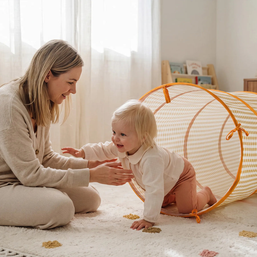 A smiling toddler crawls out of a yellow gingham Tiny Land® Adventure Play Tunnel toward a woman sitting on the floor.|Yellow