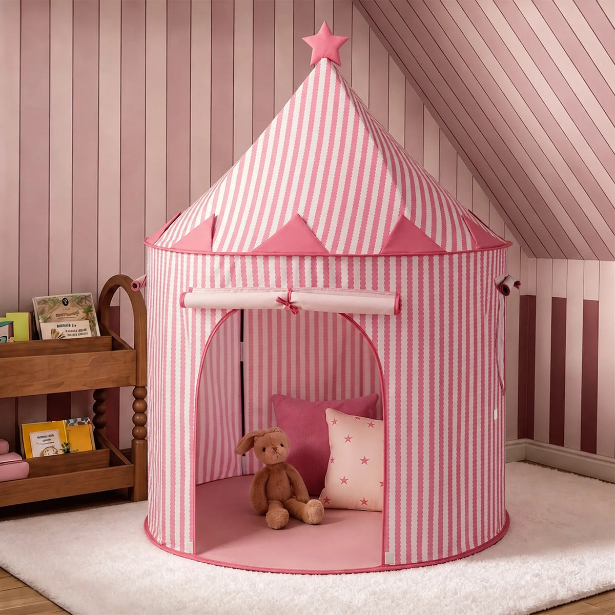 A Tiny Land® Kids Tent – Indoor Tent for Toddlers in pink and white stripes, topped with a star, stands in a pink room. Inside the tent are a brown stuffed bunny and a pink pillow decorated with stars. A wooden shelf holding books is placed nearby.|Pink