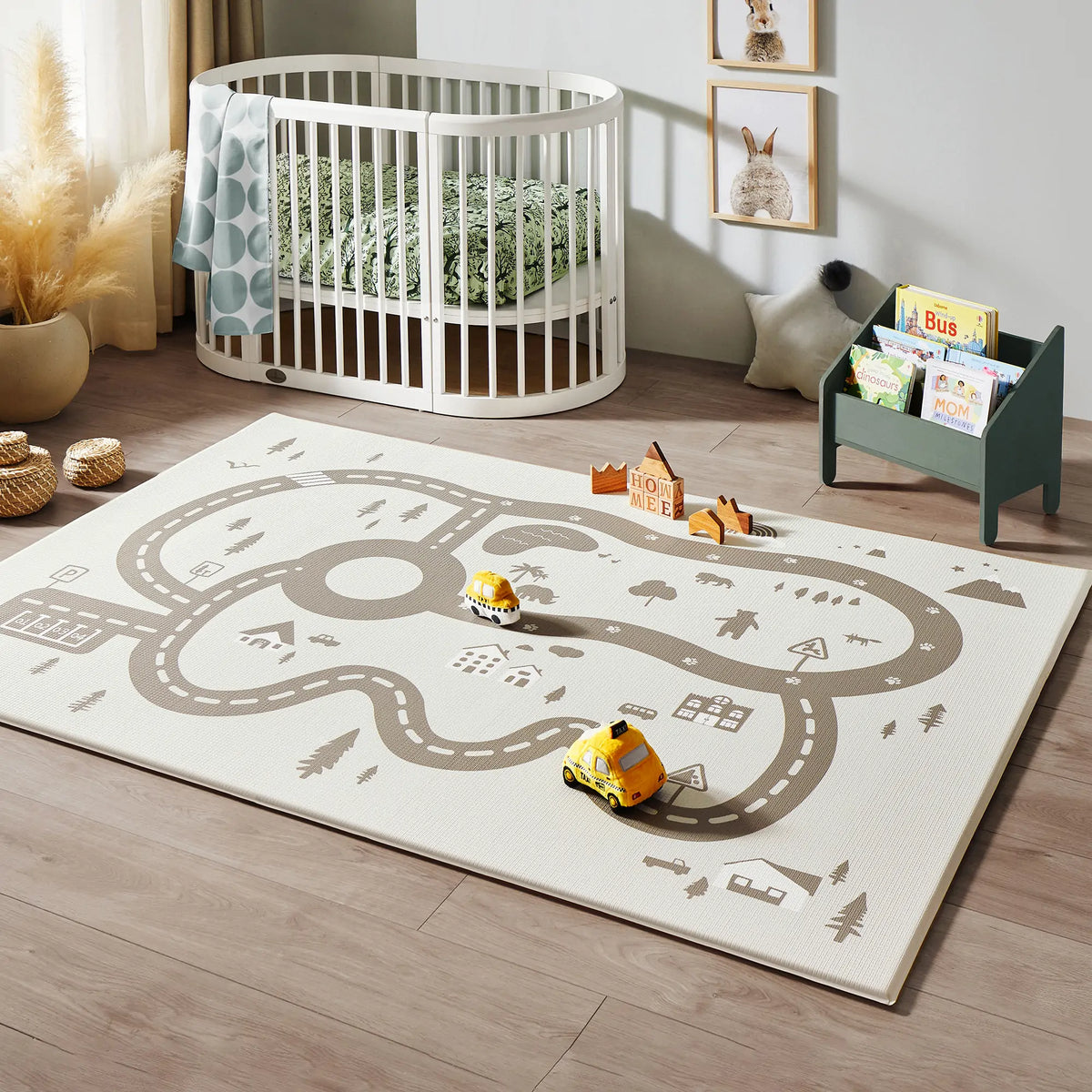 A cozy nursery features the Tiny Land Safe Explorers Playset—Natural Wood Climbing Set, the Urban Zoo Playmat with toy cars, a white oval crib with green bedding, wall art, and a small bookshelf filled with children’s books on a light wooden floor.