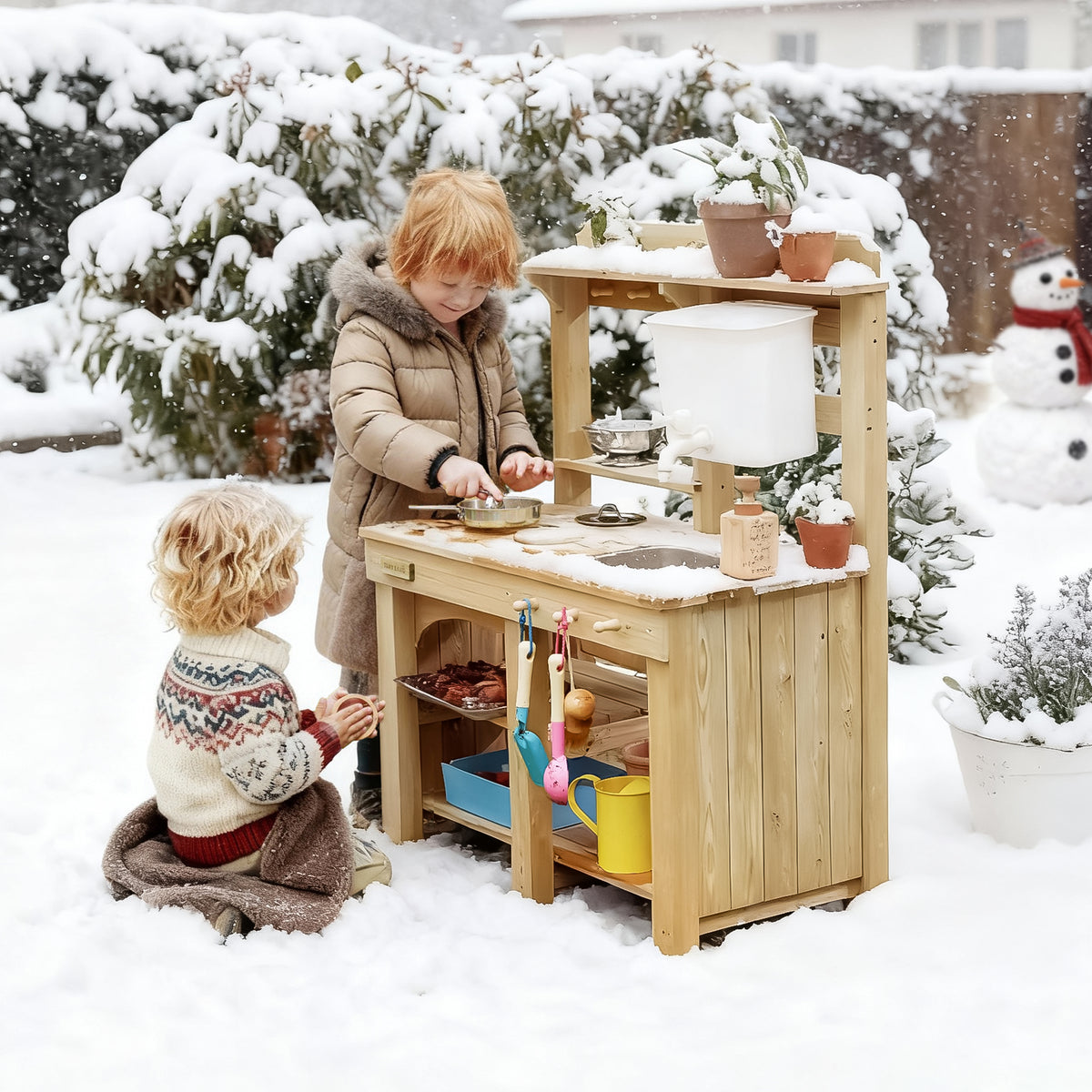 Two children play outside with the Tiny Land® Mud Kitchen - DuoPlay Outdoor Play Kitchen for Kids. One pretends to cook while the other watches, surrounded by snow-covered trees and a snowman in a wintry backyard. Brand: Tiny Land.