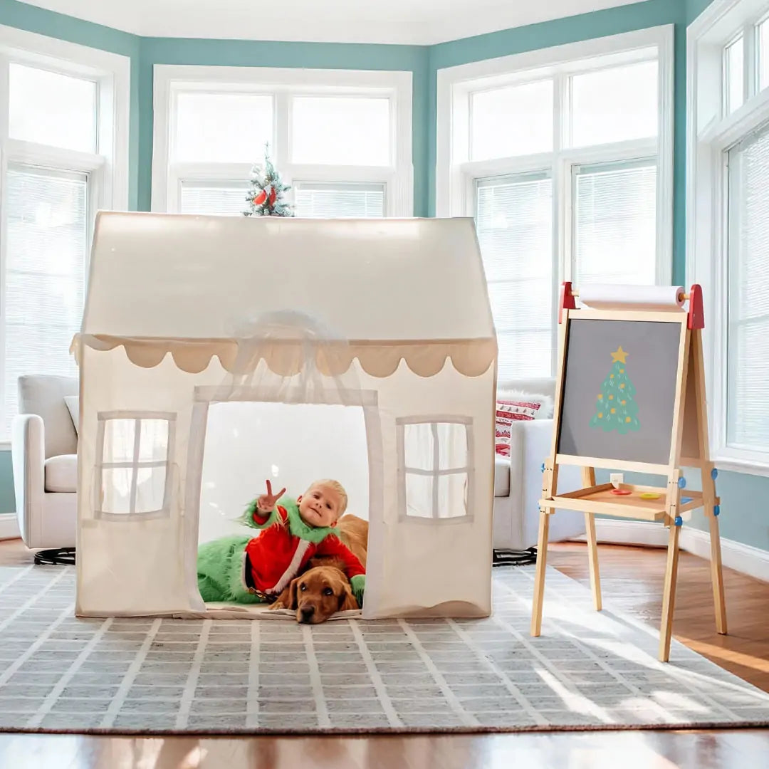 Two children in Christmas pajamas and a brown dog peek from the Tiny Land Bright Play & Art Set, featuring a cream playhouse with lights and a colorful, height-adjustable easel with holiday art, all in a festive, cozy room.