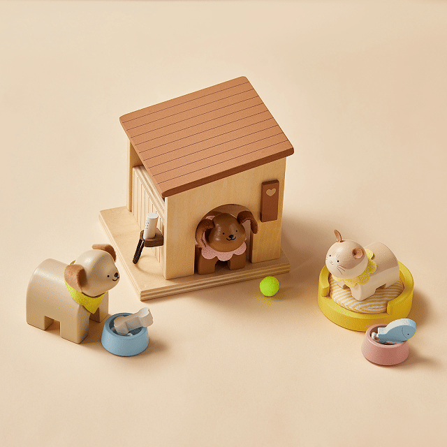 Three wooden dogs from the Tiny Land® Happy Urban Paws set by Tiny Land are arranged around a heart-decorated doghouse: one inside, one eating from a blue bowl, and one resting in a yellow bed, with toys and bowls nearby for playful fun.