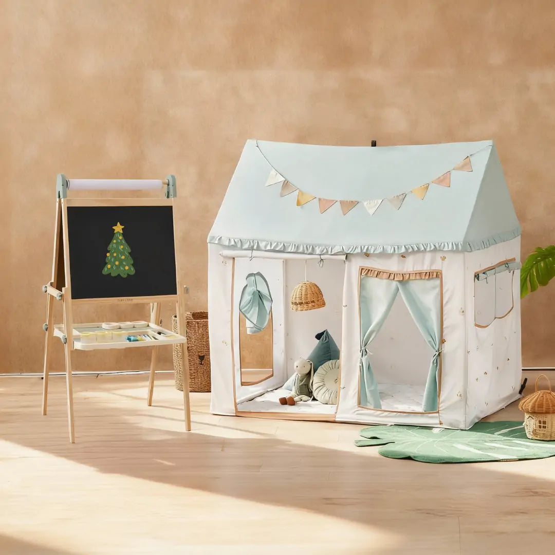 A cozy children’s playroom with the Tiny Land Nature-inspired Play & Art Set—Sage Easel + Forest Play House—plus a small basket and soft toys arranged on a wooden floor.