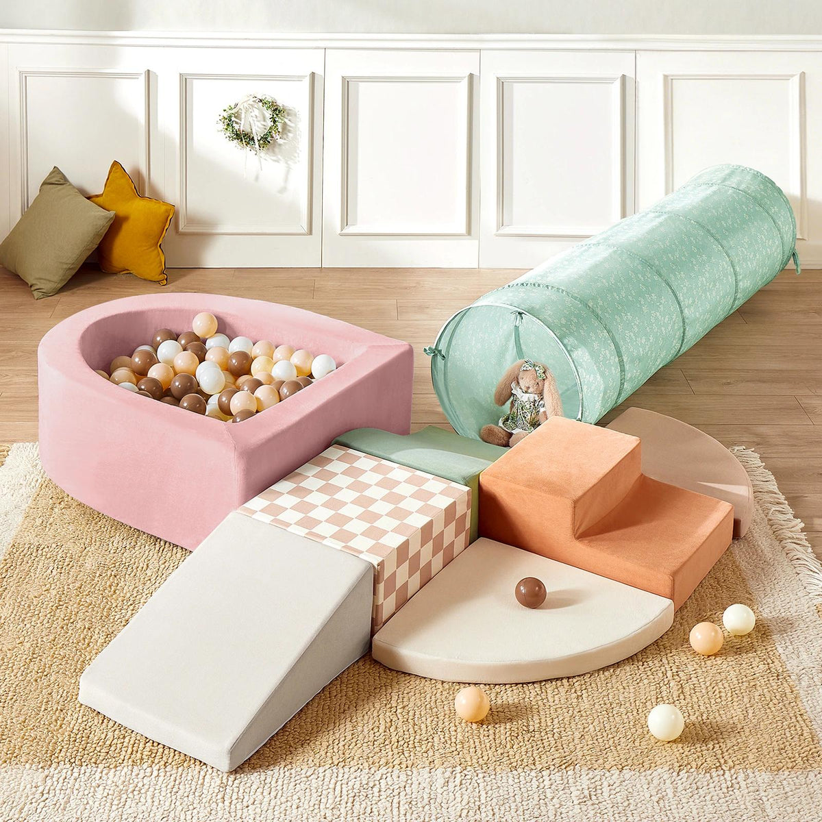 The Tiny Land® PlayNest Climbing Ball Pit Set with Tunnel by Tiny Land INC features a pastel pink foam ball pit, soft climbing blocks, gray ramp, mint green tunnel, play balls on a beige rug, and decorative pillows in the background.|pink