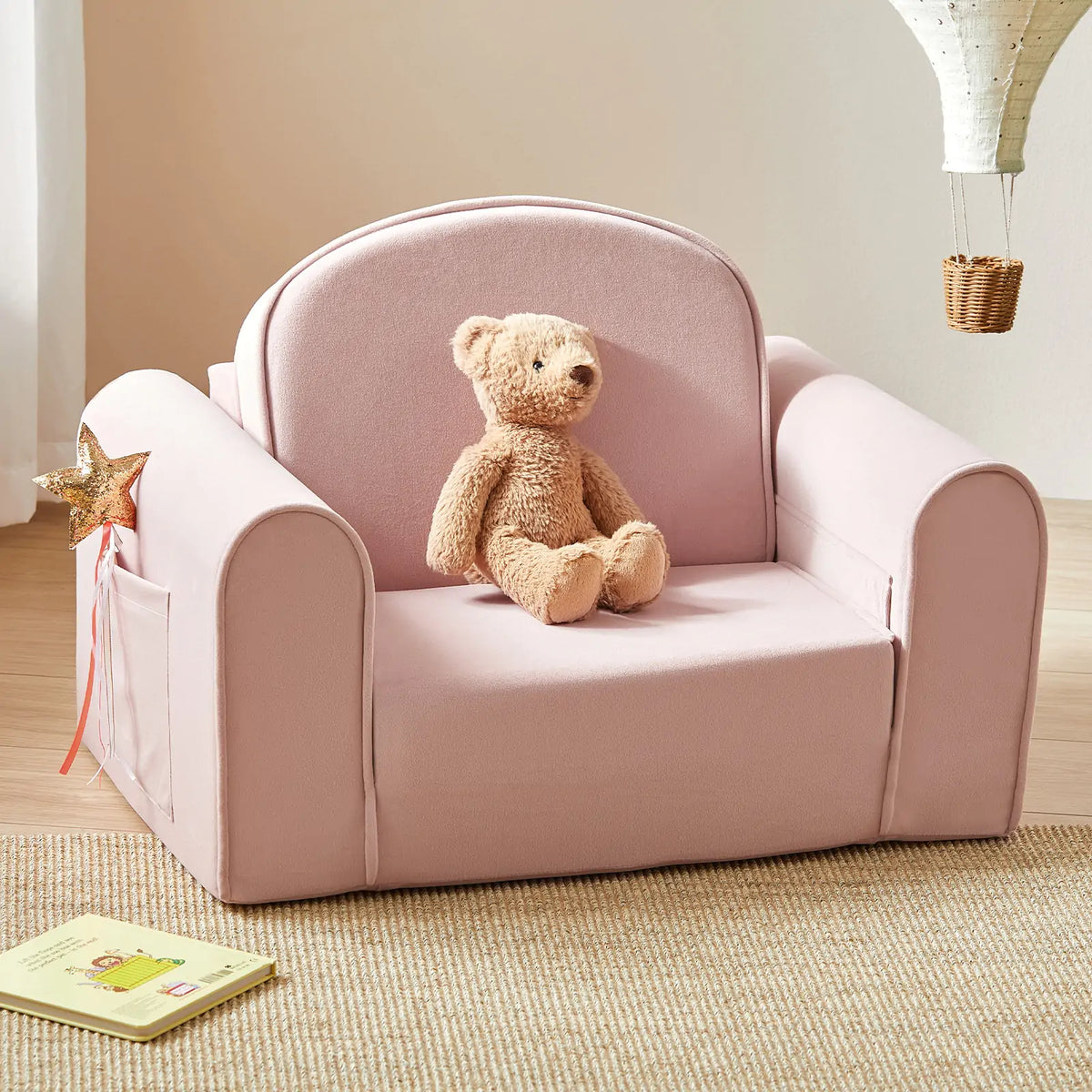 The Tiny Land® GentleSnug Kid Chair in pink adds charm to any playroom, topped with a teddy bear and featuring a pocket holding a gold-star wand. Nearby, a beige rug and paper envelope rest on the wooden floor.