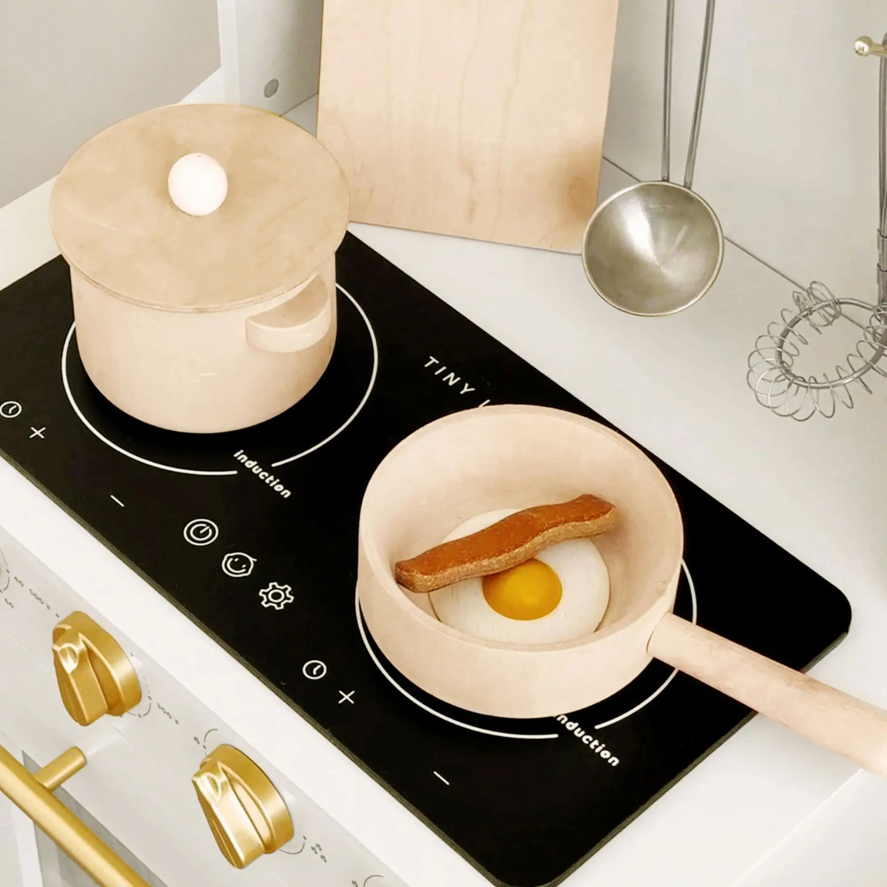 The Tiny Land® Iconic Wooden Play Kitchen features a cream finish, black stovetop, golden knobs, a wooden pot with lid, and a pan with toy egg and sausage—plus hanging utensils—for inspiring pretend cooking fun.