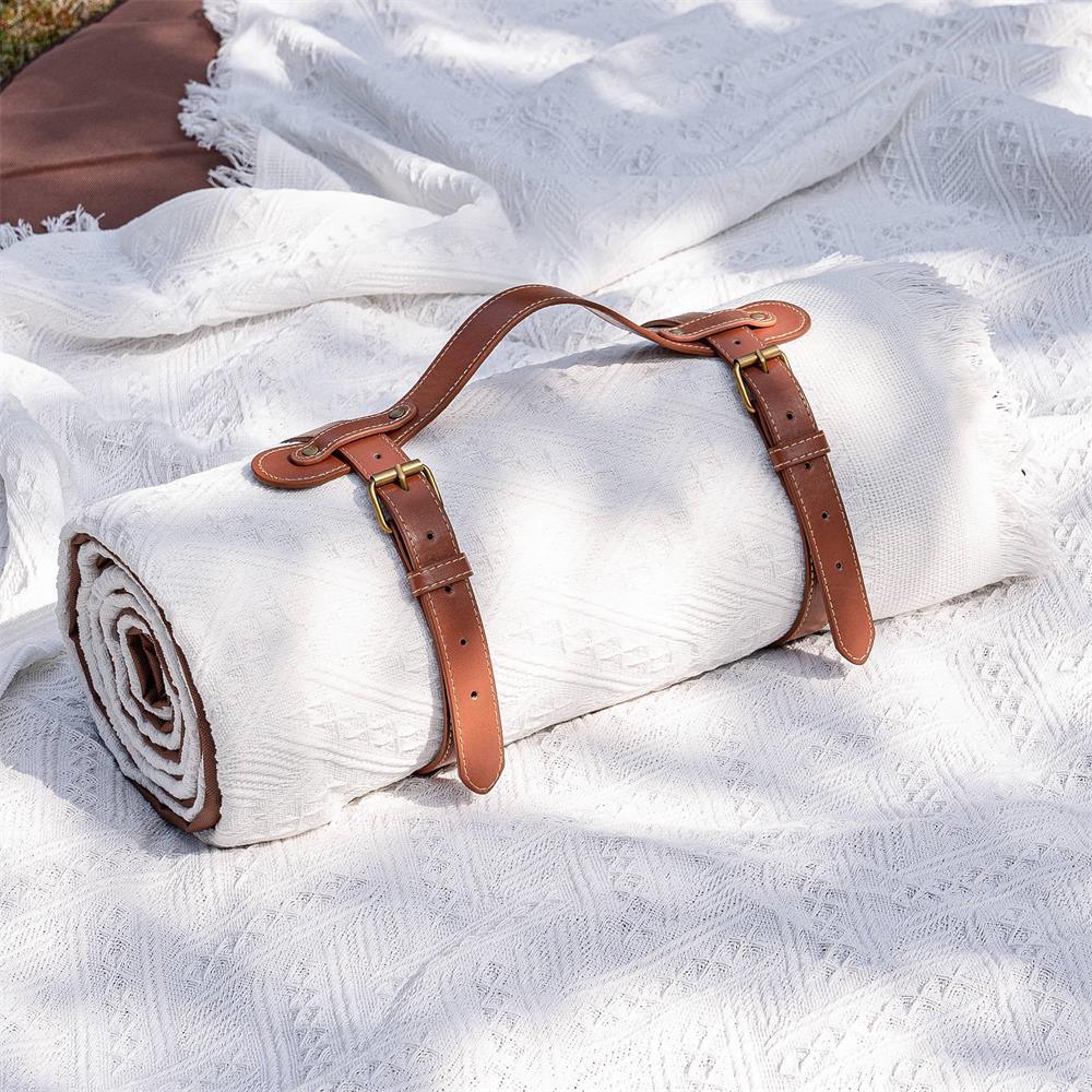 The Tiny Land® Picnic Blankets for Family by Tiny Land features a rolled-up white, textured blanket secured with brown leather straps, buckles, and a handle, resting on an unrolled matching blanket spread over grass.