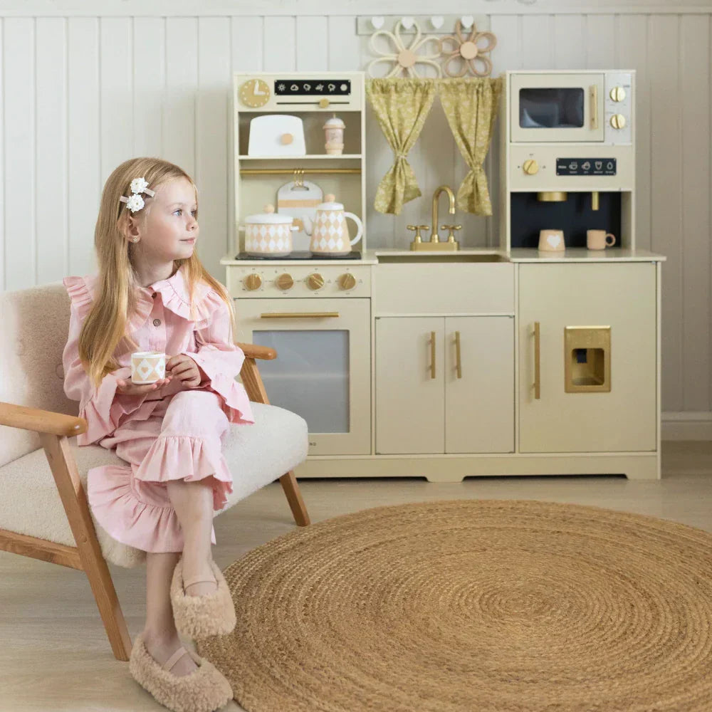 A little girl dressed in a pink outfit is sitting in front of a cream white play kitchen, holding a cup.
