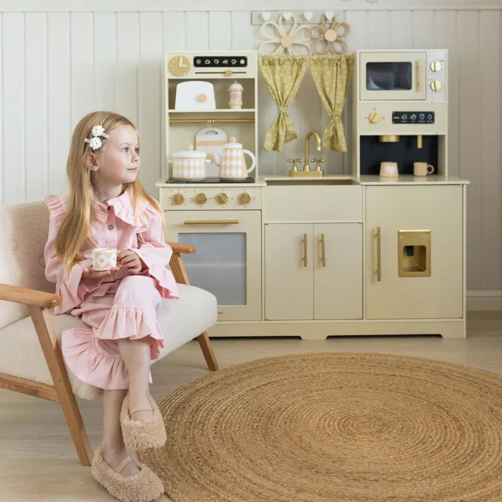 A little girl is sitting on her chair, behind her back is a beautiful play kitchen 