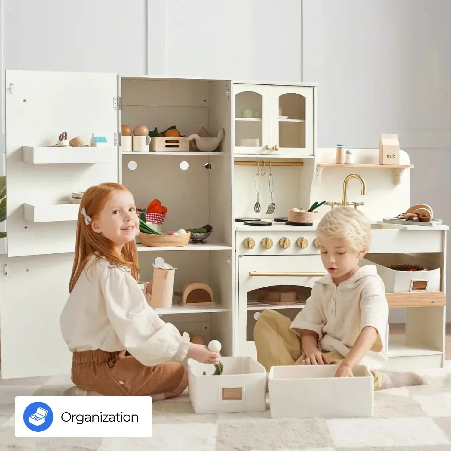 A red-haired little girl and a blonde-haired little boy are sitting on the floor of the playroom, playing pretend play games. Behind them are a cream-colored toy kitchen and a toy refrigerator.