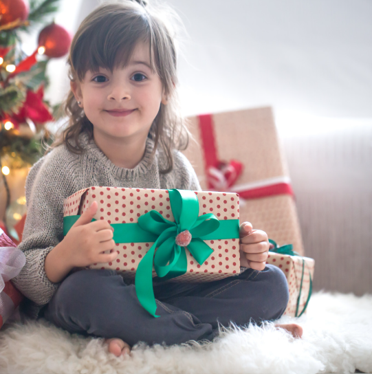 a kid is sitting on the soft mat, holding a gift box next to a Christmas tree with a happy smile 