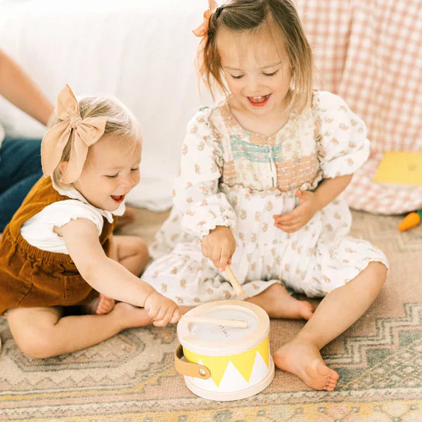 Two girls wearing matching outfits and bows on their heads sit on a blanket on the floor, happily playing a Montessori drum.