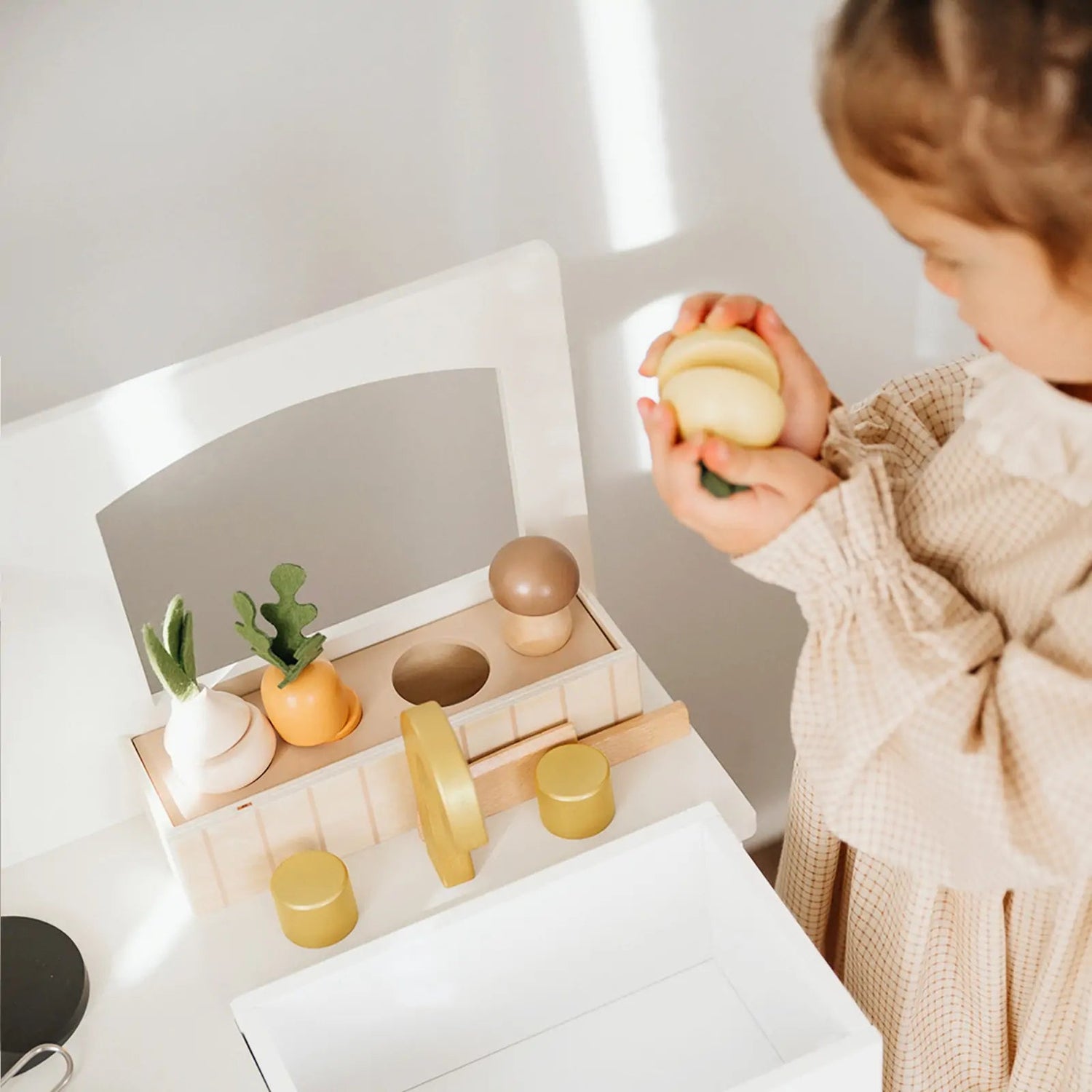 A little girl wearing a dress is standing in front of a white wooden play kitchen, playing with play food.