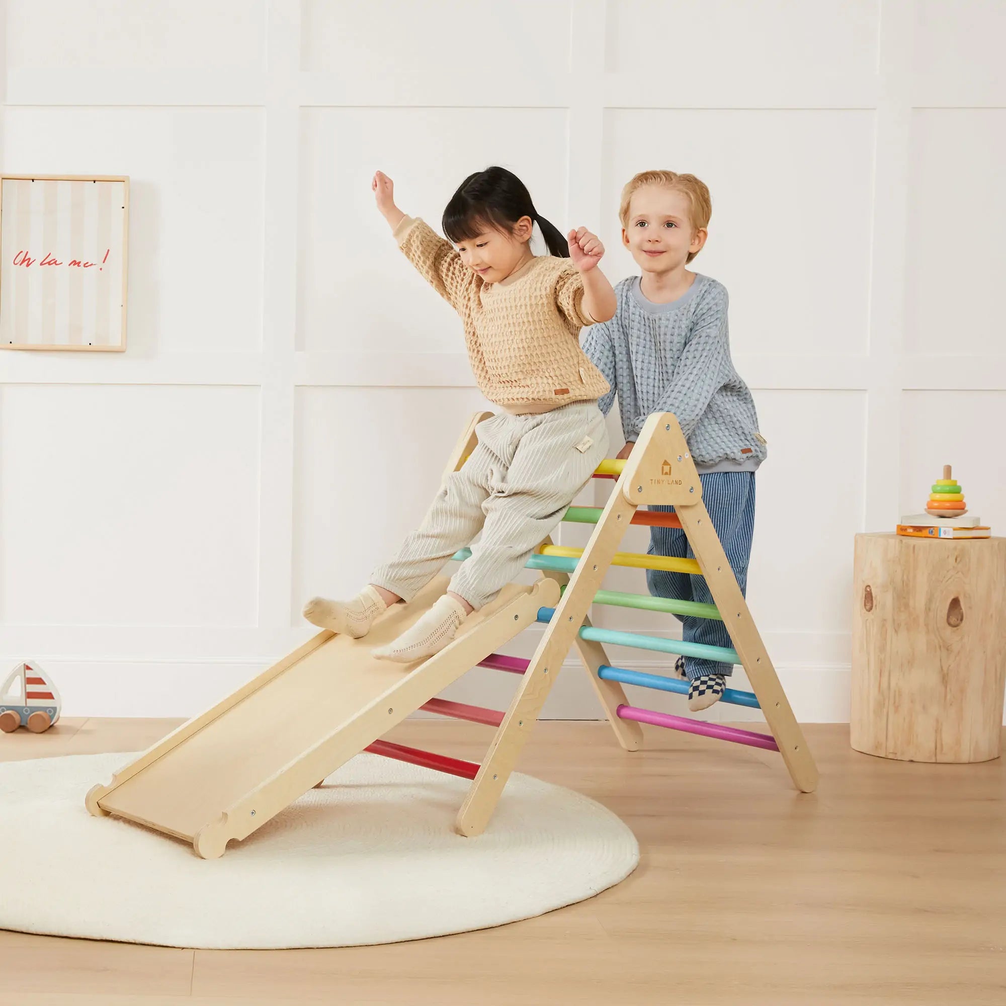 A blond little boy dressed in blue and a black-haired little girl wearing long sleeves and pants happily play on a wooden climbing structure indoors.