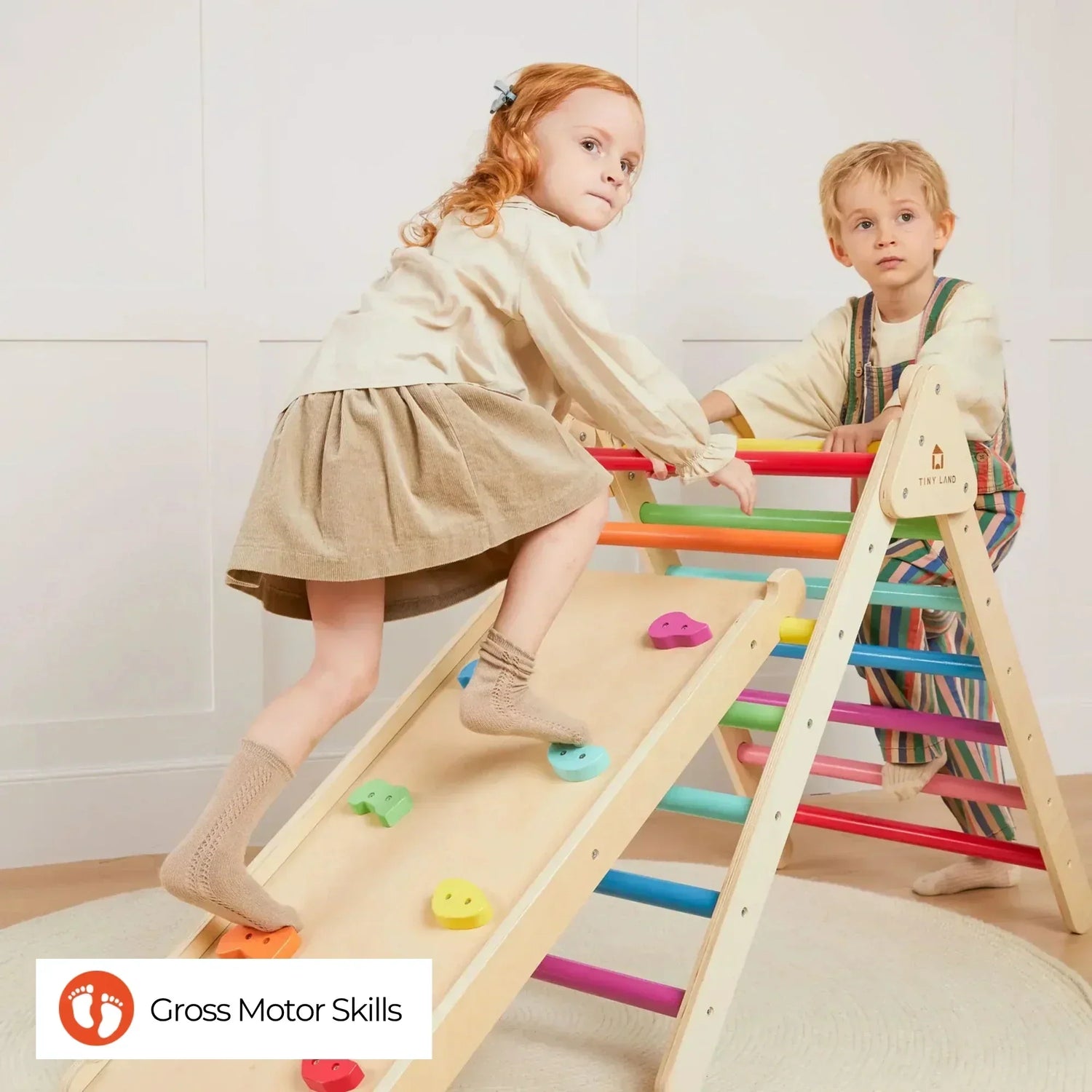 A little girl wearing a dress and a little boy wearing overalls are playing together on a rainbow-colored wooden climbing frame.