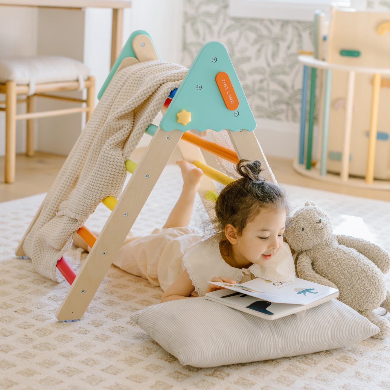 A little girl is happily reading a book while lying on a pillow beneath a triangular climbing frame.
