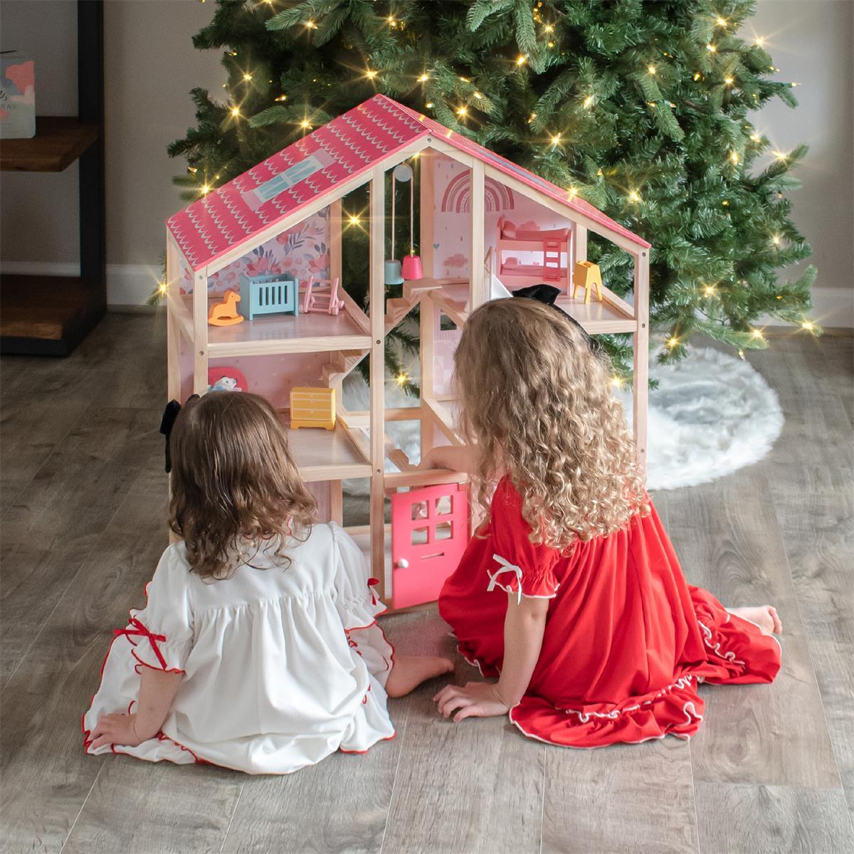 Two little girls wearing pink dresses sit by the Christmas tree, playing with a pink dollhouse.