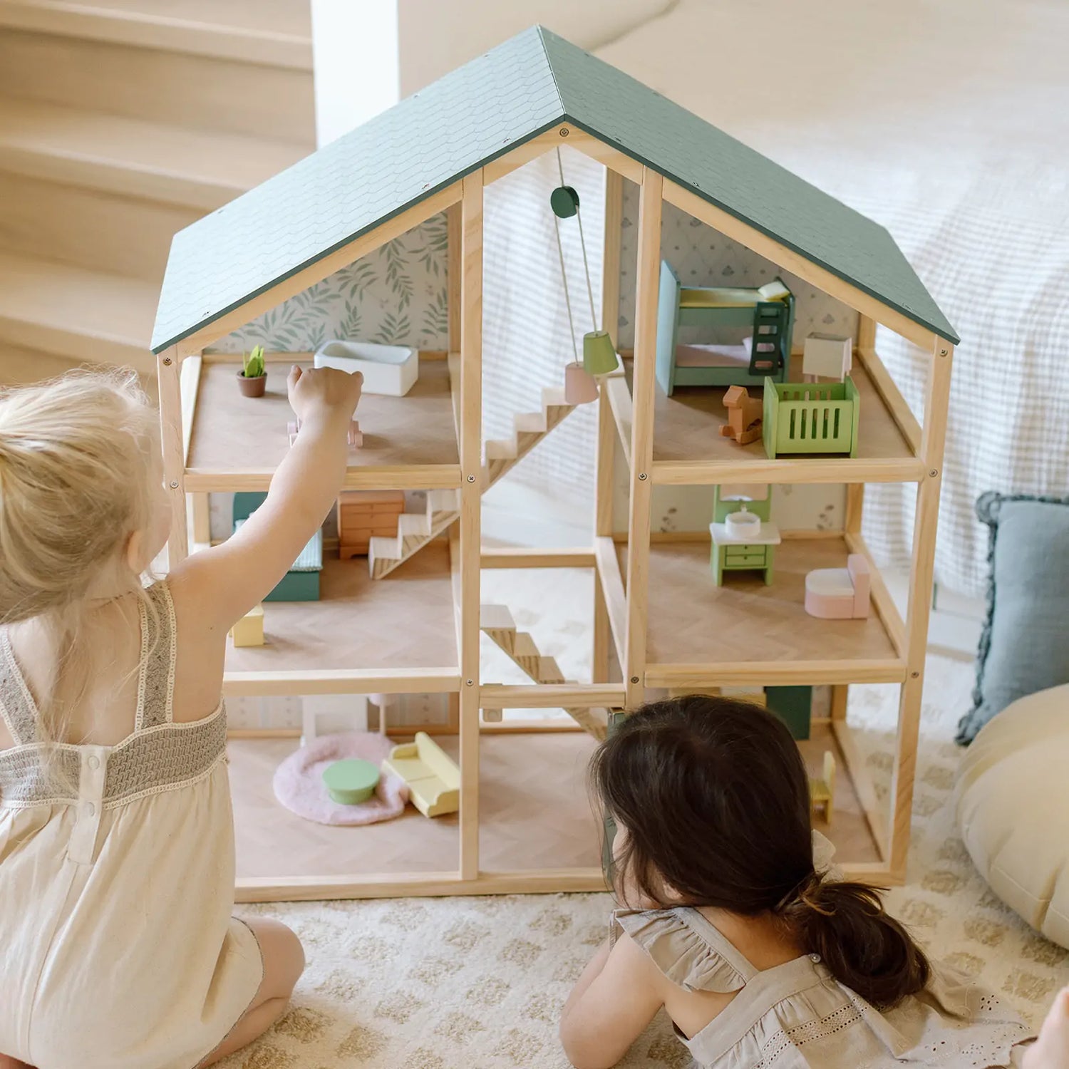 Two little girls lie on the floor playing with a green wooden dollhouse.