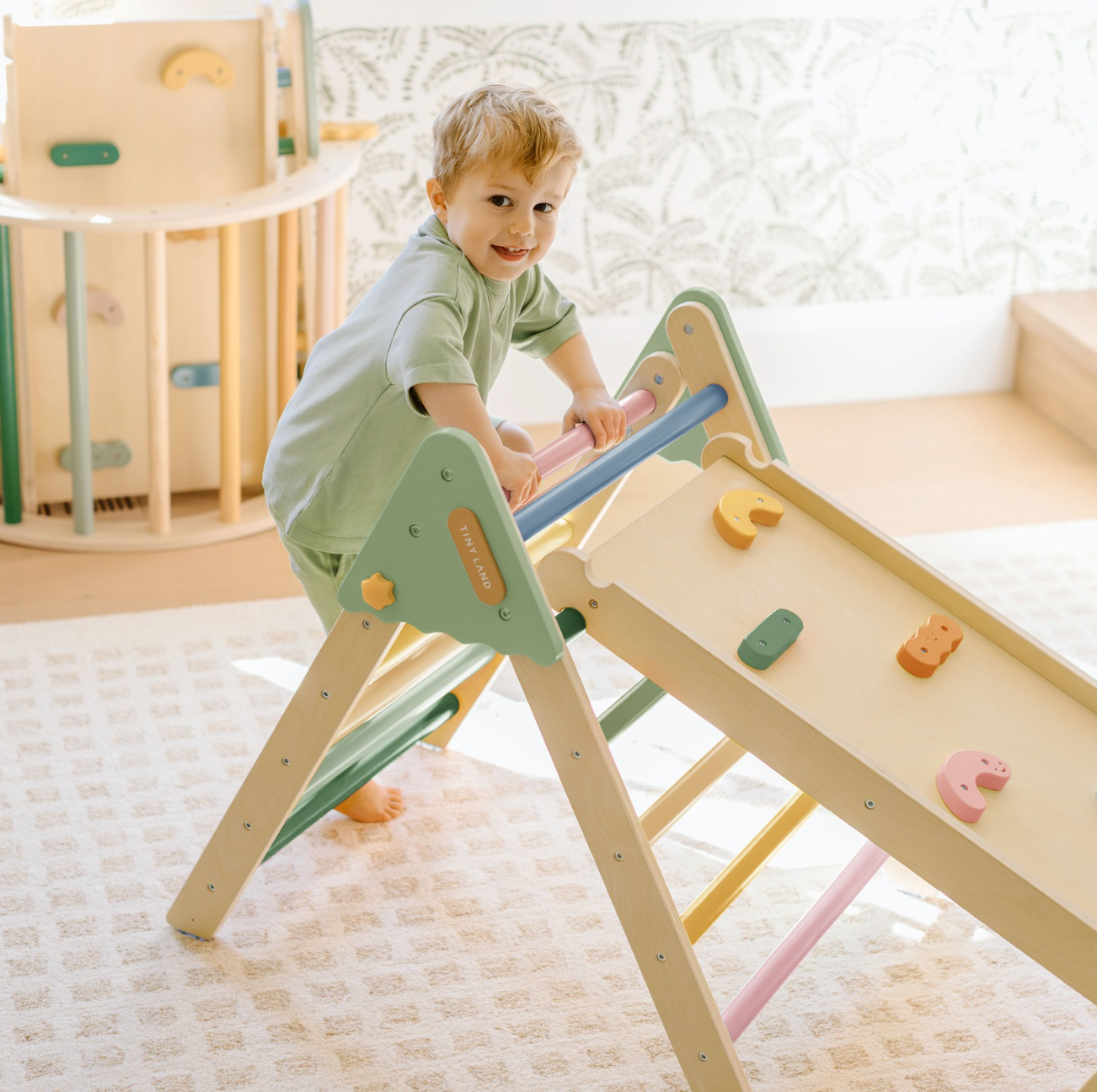A blond little boy wearing a green outfit plays on a Morandi-colored climbing set in a sun-filled playroom.