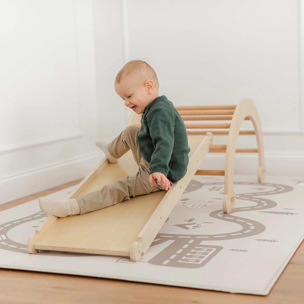 A little boy wearing a green top and khaki pants is playing on a natural wood climbing set on a play mat.