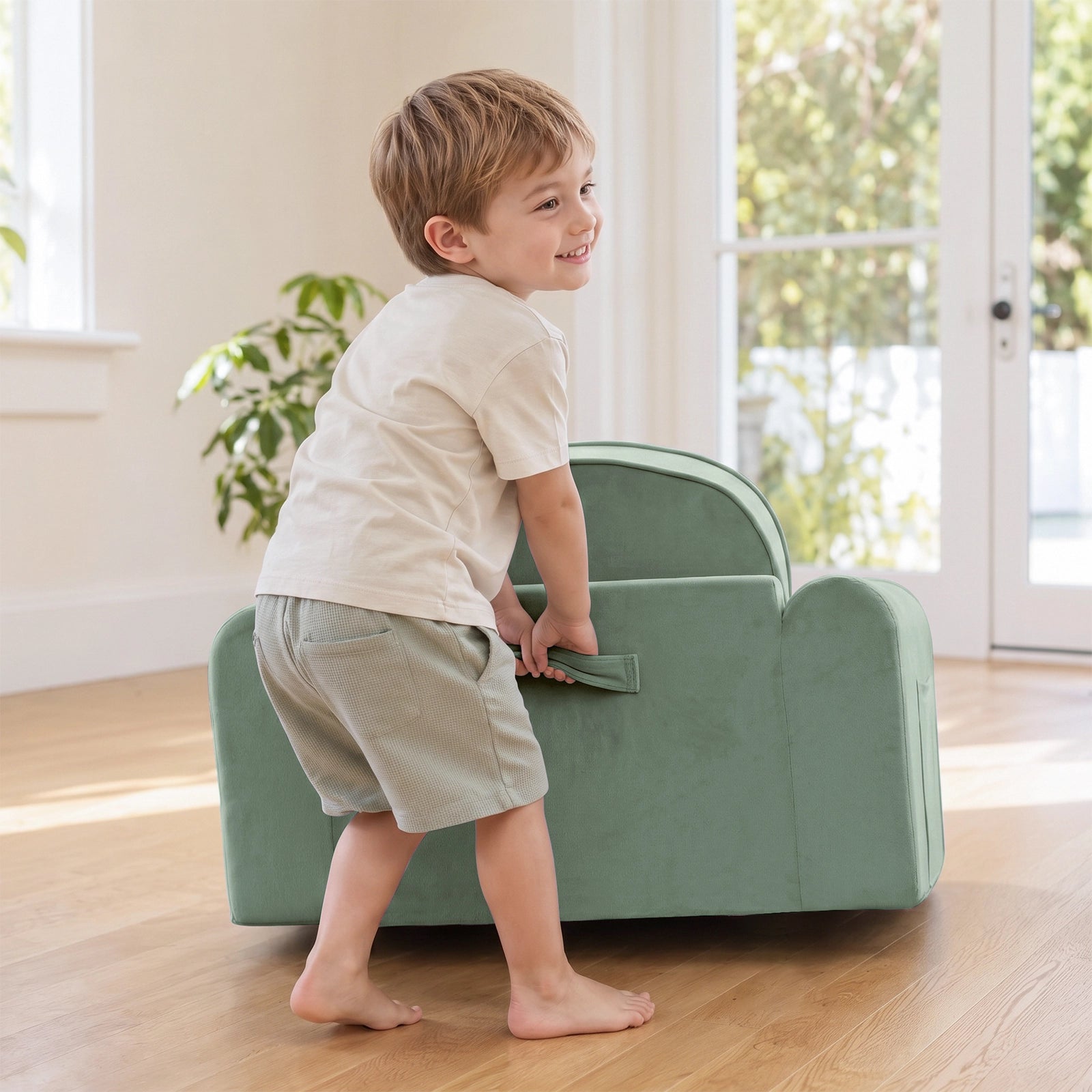 A young boy smiles while lifting a Tiny Land® GentleSnug Kids Sofa Chair by its handle in a bright, sunlit room with wooden floors and a plant in the background.|Light Green