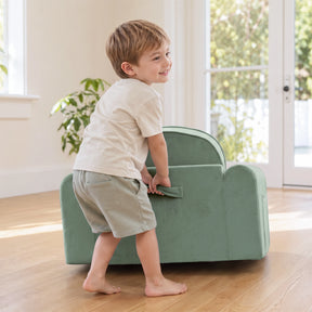 A young boy smiles while lifting a Tiny Land® GentleSnug Kids Sofa Chair by its handle in a bright, sunlit room with wooden floors and a plant in the background.|Light Green