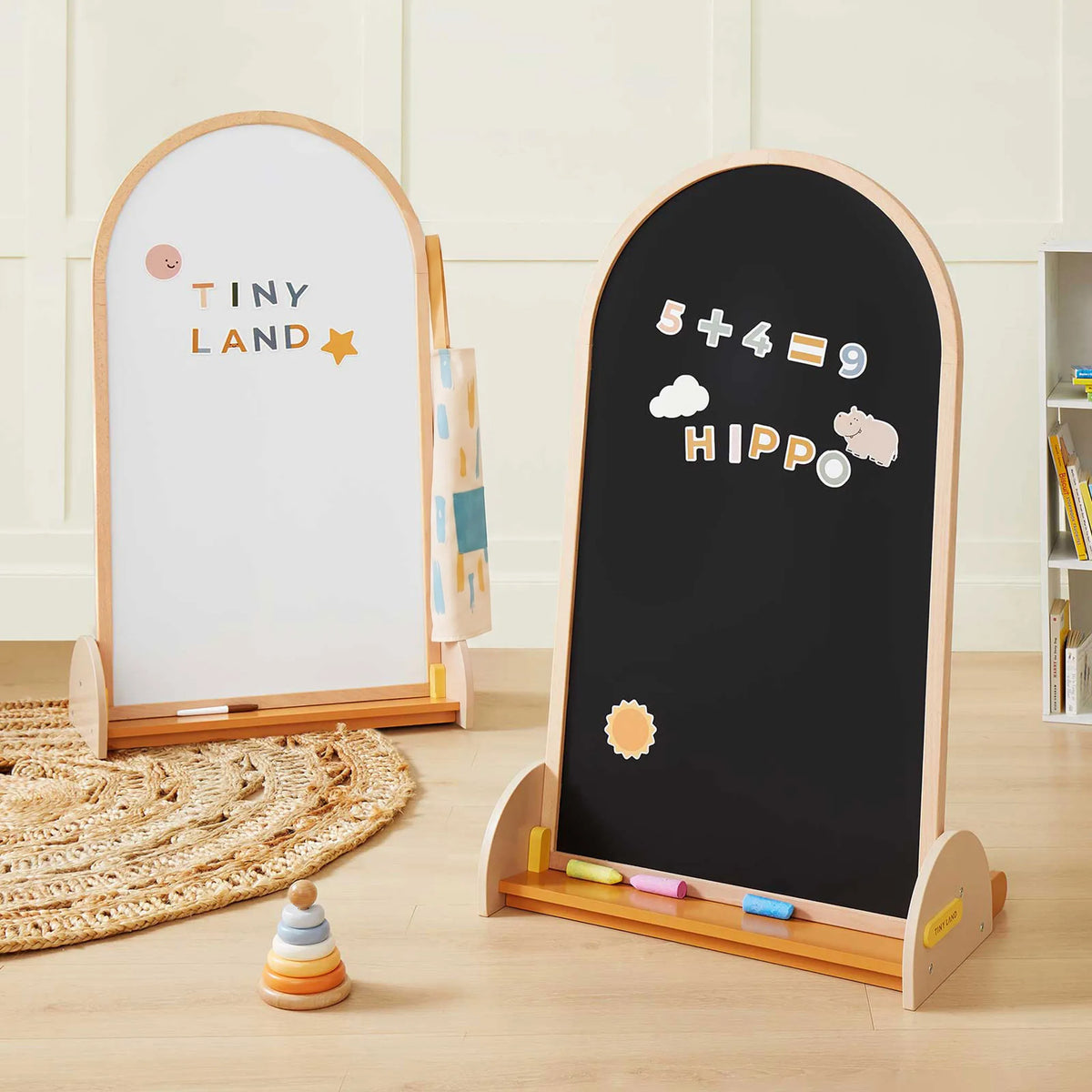 The Tiny Land® Double-Sided Arched Magnet Chalkboard by Tiny Land features a double-sided magnetic easel with a whiteboard labeled “TINY LAND” and a blackboard displaying “5 + 4 = 9” and the word “HIPPO” created with magnets. Crafted from beechwood, this kids’ art board comes with chalk and toys placed nearby on the wooden floor, making it a versatile chalkboard whiteboard set for children.